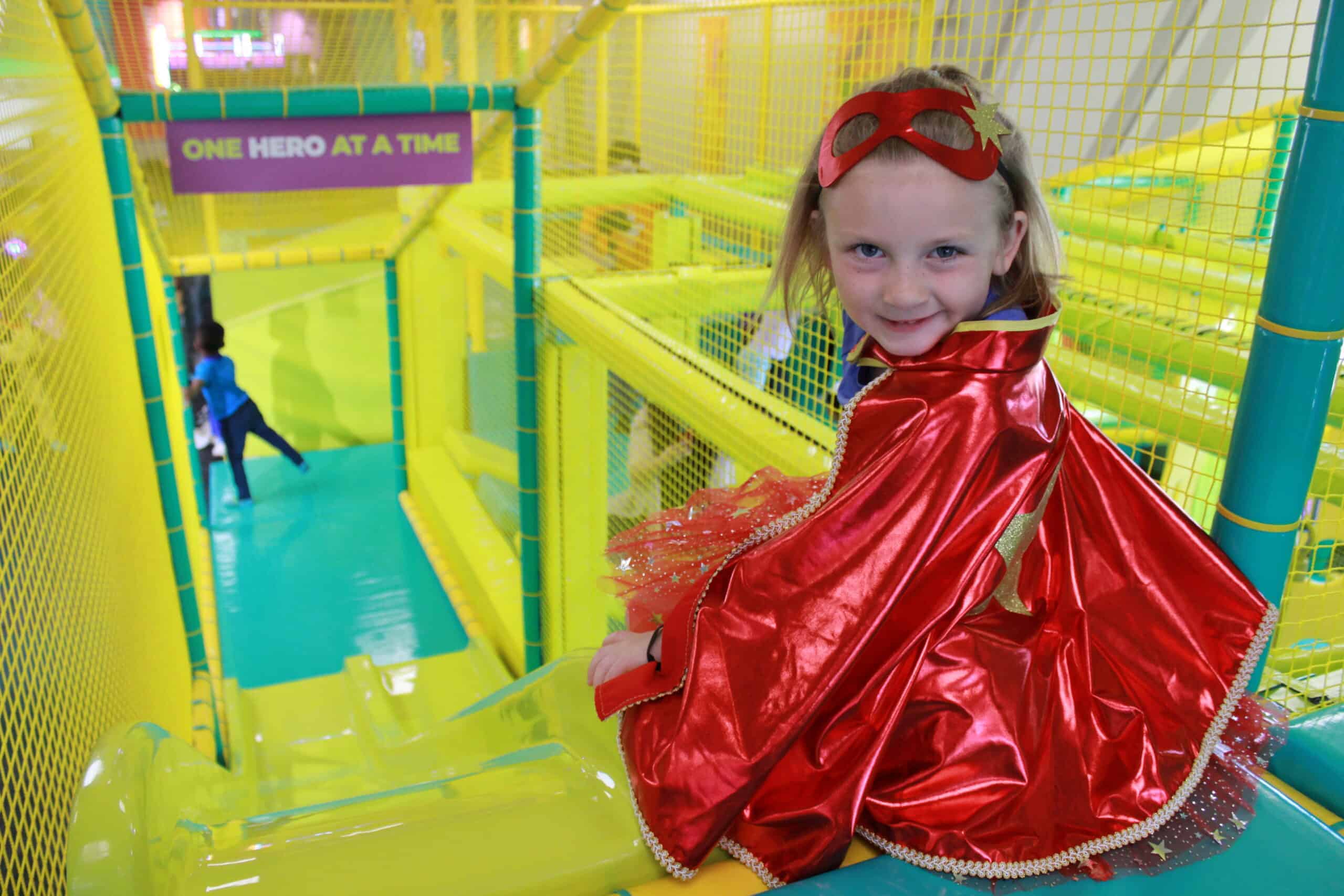 A young child in a costume cape at the top of a slide in the toddler zone