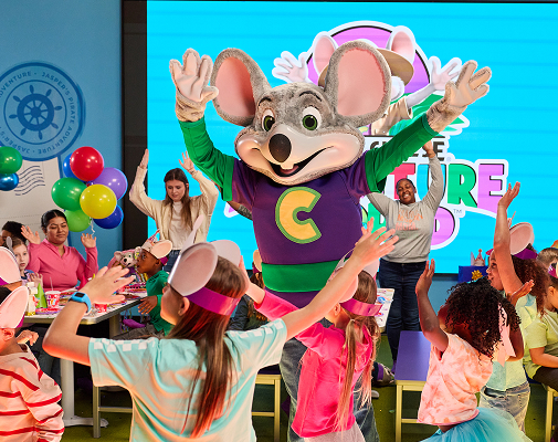 Adventure Chuck E. leading a live show on stage while kids cheer from their party table